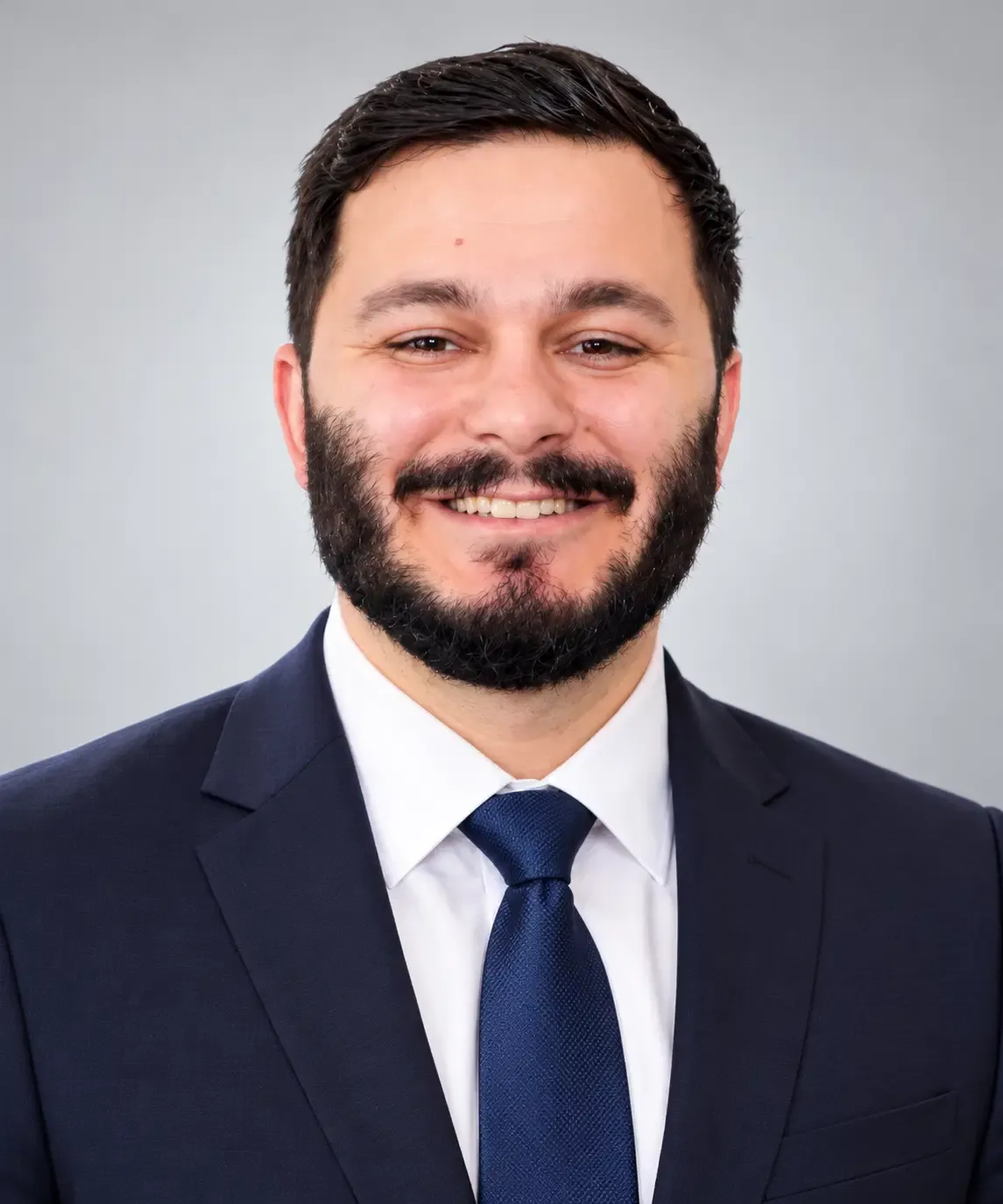 Portrait of Nick Contarino, a smiling man with dark hair and a full beard in a navy suit, white shirt, and dark tie.